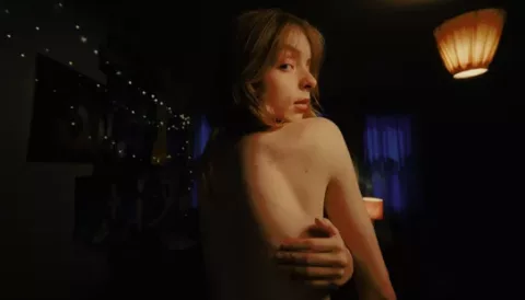 A woman in a bedroom with her back to camera, looking over her shoulder as her hand is reached around her back as she is checking her body