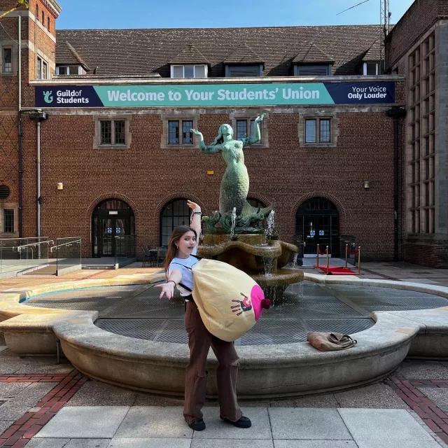 A woman stood in front of a fountain with her hands in the air, wearing a boob suit